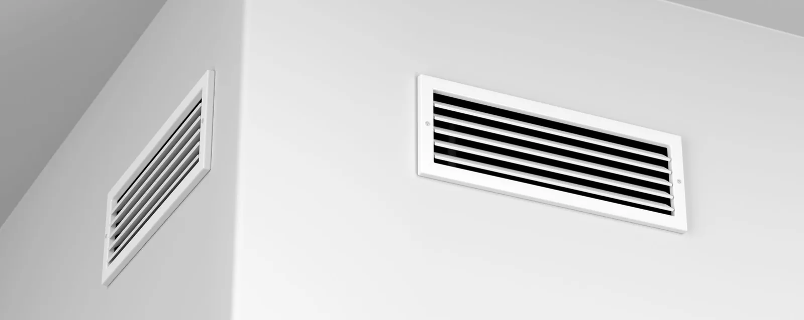 Deux grilles d'aération rectangulaires blanches sur un mur gris clair.