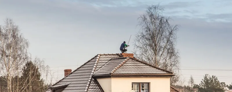 Personne sur une cheminée de toit ; toit enneigé, arbres dénudés, ciel couvert.