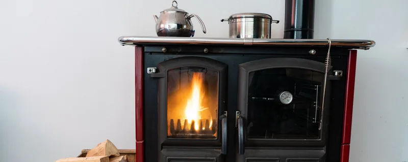 Un poêle à bois allumé avec une théière et une casserole dessus ; du bois de chauffage empilé en dessous.