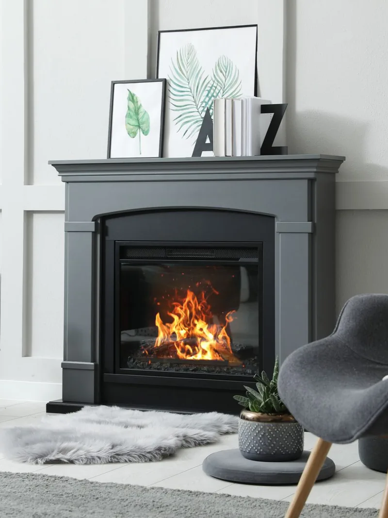 Cheminée grise aux flammes crépitantes, ornée d'œuvres d'art et de livres. Un fauteuil gris se trouve à proximité.