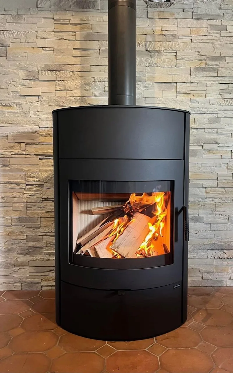 Cheminée cylindrique noire avec des bûches qui brûlent, près d'une pile de bois de chauffage.